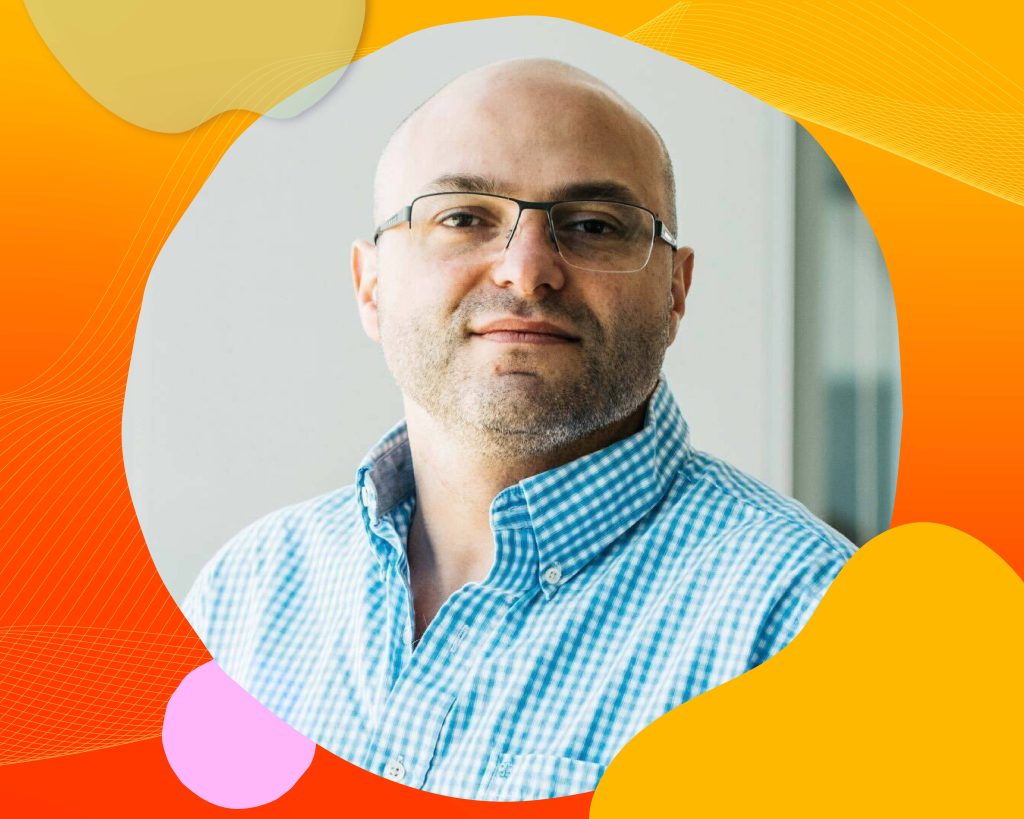 Joseph is wearing a blue checkered collared shirt. He's wearing glasses while looking at the camera smiling.