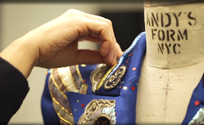 Das Bild zeigt eine Nahaufnahme einer Hand einer Schneiderin, die gerade Pailetten auf eine blaue Jacke von Marc Jacobs näht.