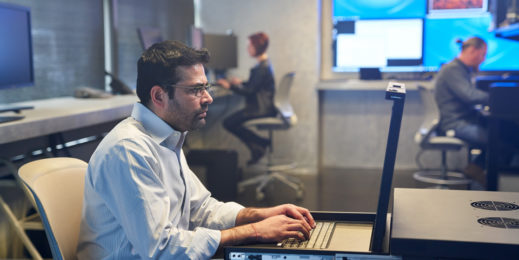 Mann mit Kragenhemd, der auf einer Serverstation in einem Sicherheitsraum arbeitet. Beschäftigte und große Monitore sind im Hintergrund zu sehen.