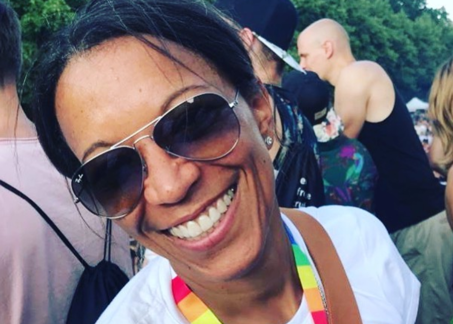 Porträtfoto von Janina Kugel, lachend mit einer Sonnenbrille und einem Band in Regenbogenfarben um den Hals