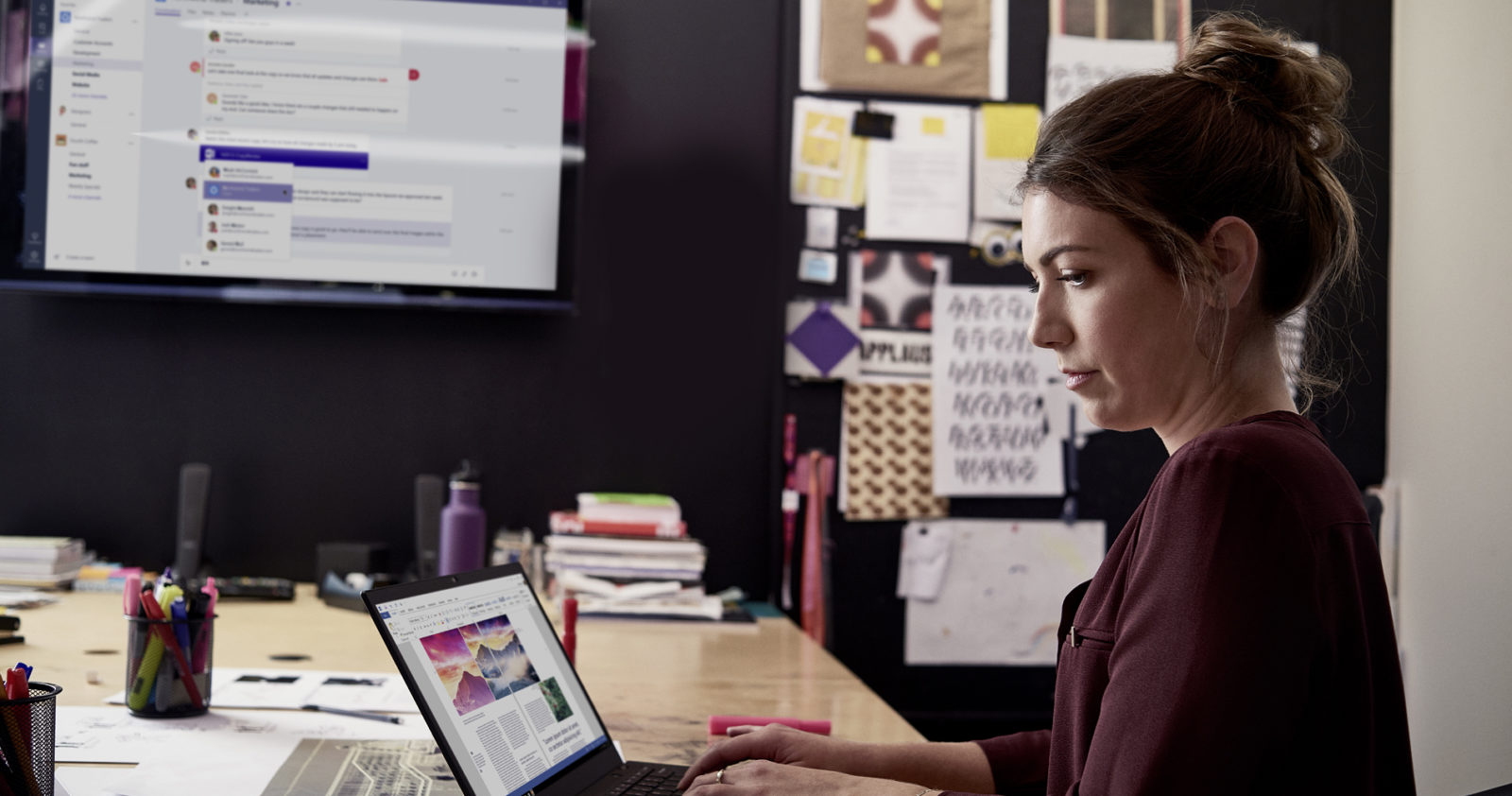 Eine Frau sitzt in einem Konferenzraum und arbeitet an ihrem Laptop. Im Hintergrund befindet sich ein Monitor, auf dem Teams gezeigt wird.