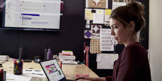 Eine Frau sitzt in einem Konferenzraum und arbeitet an ihrem Laptop. Im Hintergrund befindet sich ein Monitor, auf dem Teams gezeigt wird.