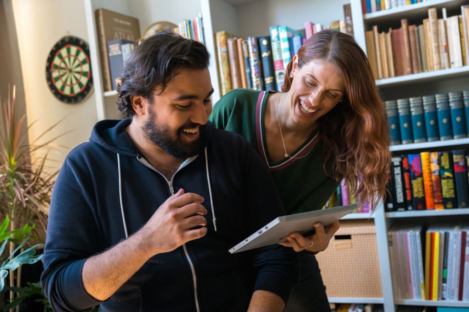 Zwei Personen stehen nah beinander im heimischen Wohnzimmer, sie lachen und schauen auf ein Endgerät in ihrer Hand. Im Hintergrund ein Bücherreal und eine Dart-Scheibe.