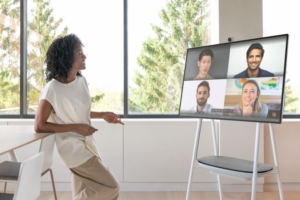 Eine Frau steht in einem Konferenzraum vor einem Surface Hub und spricht zu ihren Kolleg*innen in einer Teams-Besprechung.