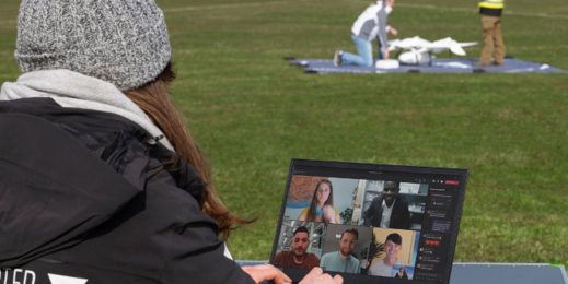 Im Vordergrund eine Frau mit Mütze, vor ihr steht ein Laptop, auf dem eine Teams Besprechung läuft. Im Hintergrund zwei Personen, die eine Drohne für den Flug vorbereiten.