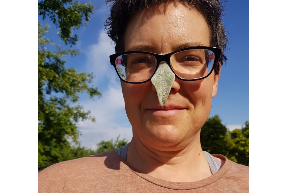 Das Gesicht eines Menschen mit kurzen dunklen Haaren und einer Brille, auf der Nase ein grünes Blatt. Die Person steht draussen in der Sonne, im Hintergrund blauer Himmel und grüne Bäume.