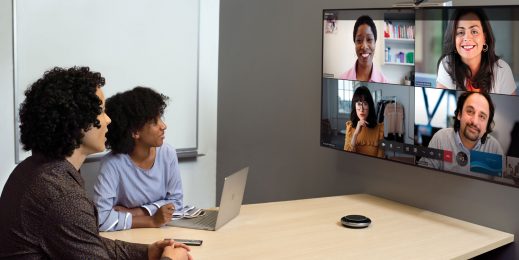 Zwei Menschen sitzen in einem kleinen Konferenzraum, sie blicken auf einen Bildschirm an der Wand, auf dem vier Teilnehmer*innen der Teams-Besprechung zu sehen sind.