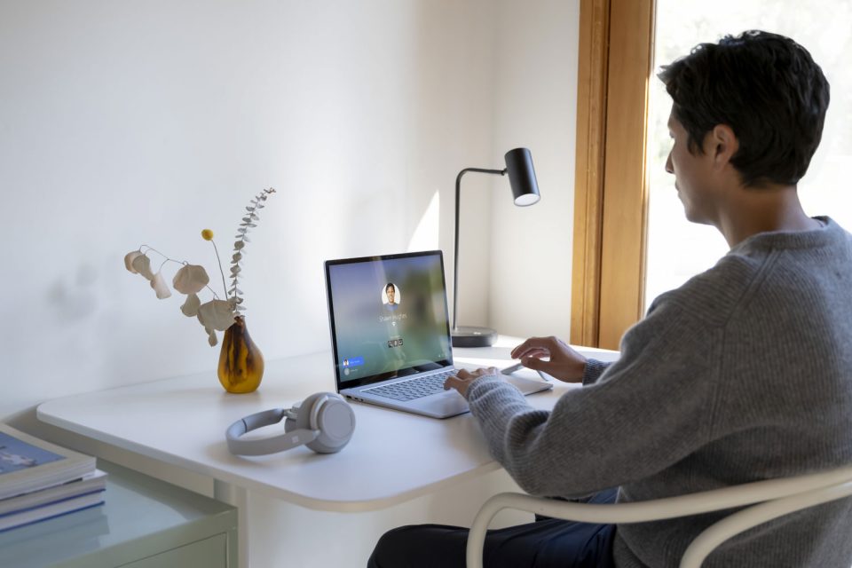 Eine Person sitzt an einem Schreibtisch, der Schreibtisch steht an einer weißen Wand. Auf dem Tisch steht ein Laptop, auf dem sich die Person gerade anmeldet.