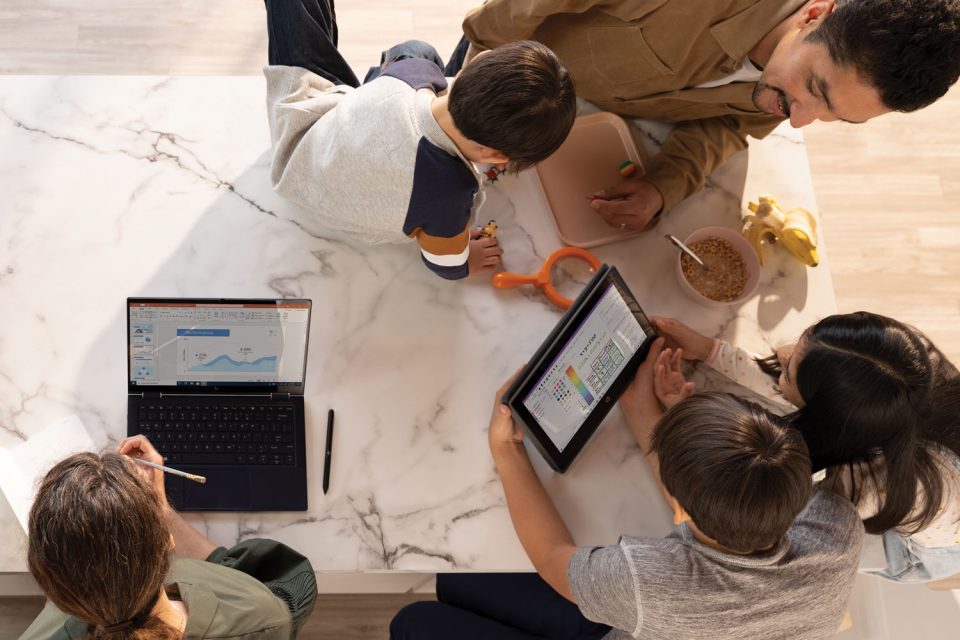 Eine Familie arbeitet und lernt aus der Ferne an ihrem Küchentisch. Die Mutter hat eine PowerPoint-Datei auf einem HP Elite Dragonfly geöffnet. Der Vater und die beiden älteren Kinder sehen sich OneNote Math Assistant auf einem HP Probook x360 an.