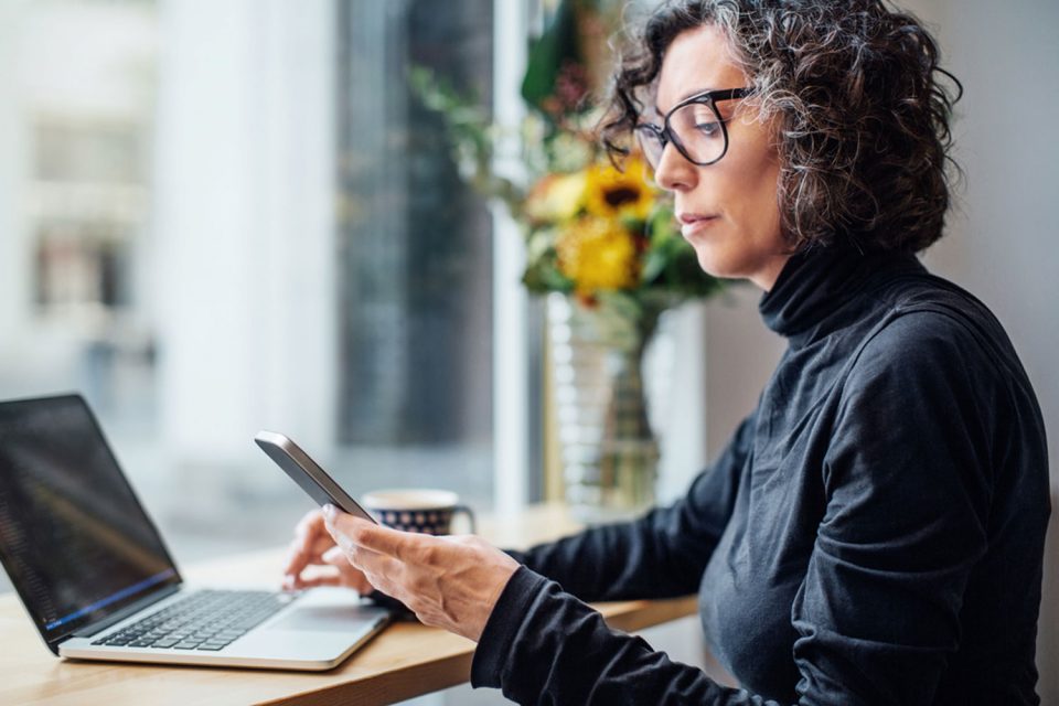 Eine Person sitzt in einem Café, sie trägt eine Brille. Vor ihr auf dem Tisch steht ein Laptop, in der Hand hält sie ein Smartphone.
