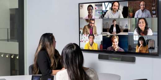 Zu sehen sind zwei Personen während eines Video-Calls. Sie befinden sich im Büro und blicken auf einen Bildschirm an der Wand, auf dem weitere Teilnehmer*innen des Meetings zu sehen sind.