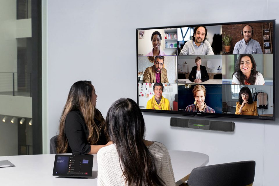 Zu sehen sind zwei Personen während eines Video-Calls. Sie befinden sich im Büro und blicken auf einen Bildschirm an der Wand, auf dem weitere Teilnehmer*innen des Meetings zu sehen sind.