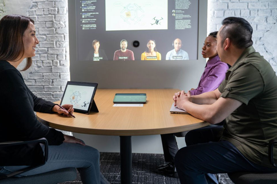 Drei Personen sitzen in einem Meeting-Raum an einem runden Tisch. Auf Augenhöhe sind ihnen Teilnehmer*innen auf Teams zugeschaltet.