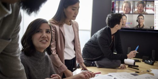 Eine Arbeitsgruppe aus drei Frauen hält ein hybrides Meeting ab. Auf dem Bildschirm im Hintergrund sind die Kolleg*innen zugeschaltet.
