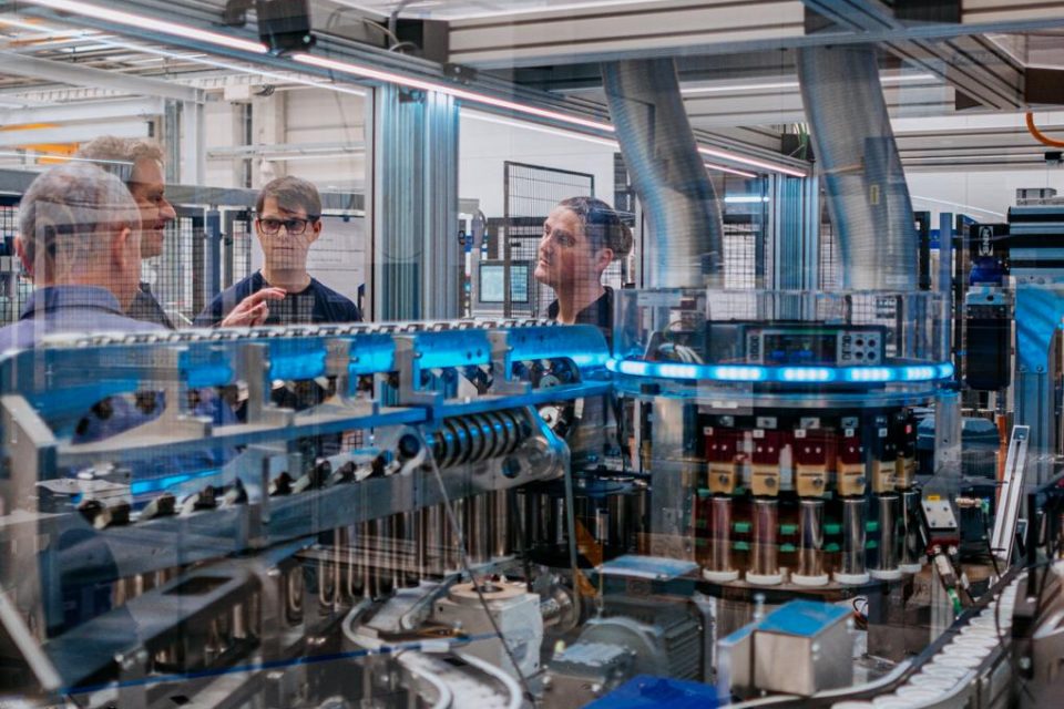 Zwei Männer mit dem Rücken zur Kamera und zwei Männer, die der Kamera zugewandt sind, wie sie durch das Glas einer komplexen Maschine zu sehen sind.