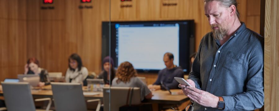 Hombre con barba mira su teléfono con el fondo de una sala de conferencias borrosa de personas que trabajan.