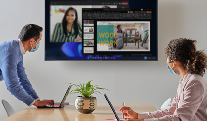 Dos personas trabajan en una sala de juntas con sus laptops y miran una pantalla