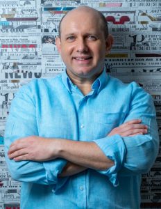 Un hombre con camisa azul y brazos cruzados sobre el pecho, posa para la cámara frente a una pared colorida