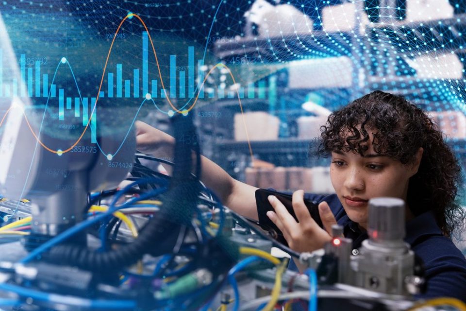 Mujer observa lo que hay en su dispositivo móvil mientras trabaja en un entorno con muchos alambres y cables. En la foto se superponen imágenes con gráficos y números.