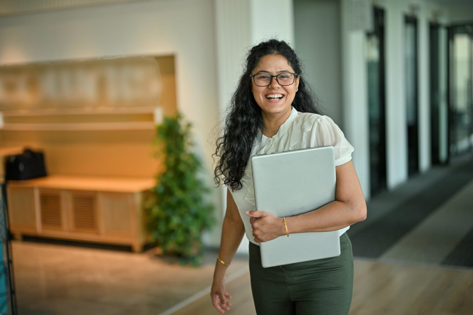 Mujer camina en un pasillo de oficinas con una laptop en la mano