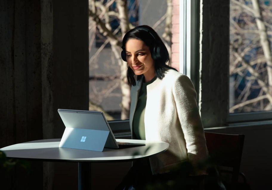 Mujer sentada en una mesa usa audífonos y observa una Surface
