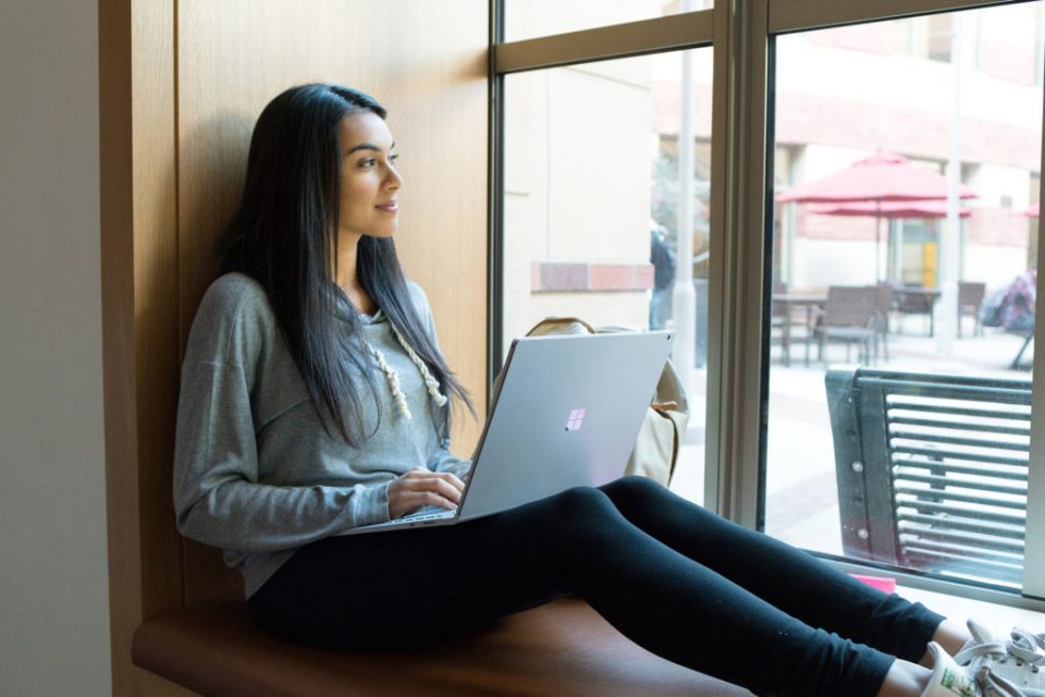 Una mujer sentada frente a una ventana con una laptop sobre sus piernas