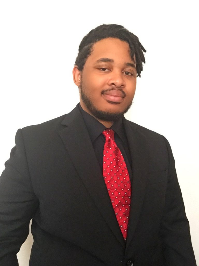 homem em pé de terno e gravata vermelha sorrindo para foto