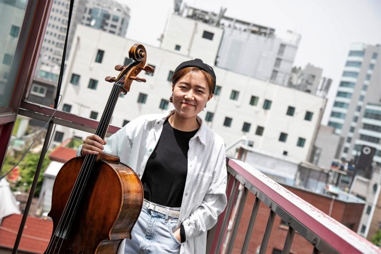 mulher em pé com uma mão no bolso da calça jeans e outro segurando violão cello