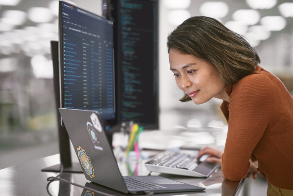 Mulher olha para tela de notebook e ao lado há um computador