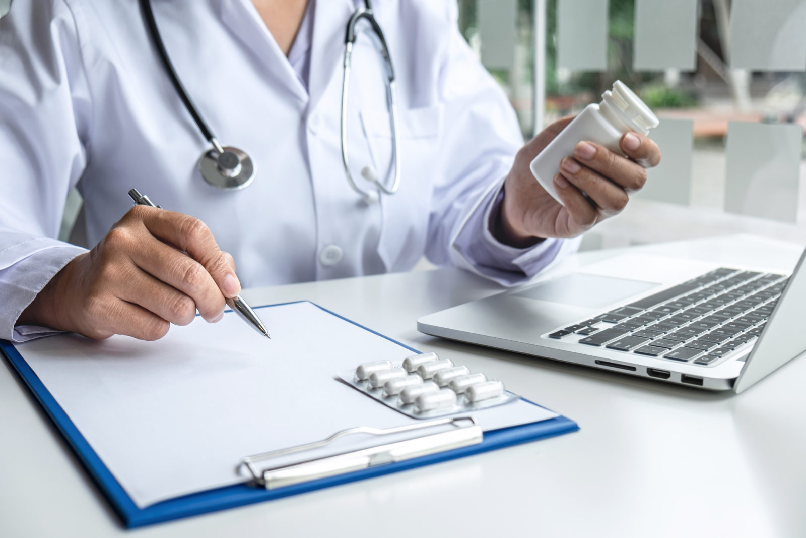 A doctor holds a pill bottle in front of a laptop and writes on a clipboard