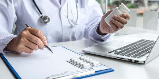 A doctor holds a pill bottle in front of a laptop and writes on a clipboard