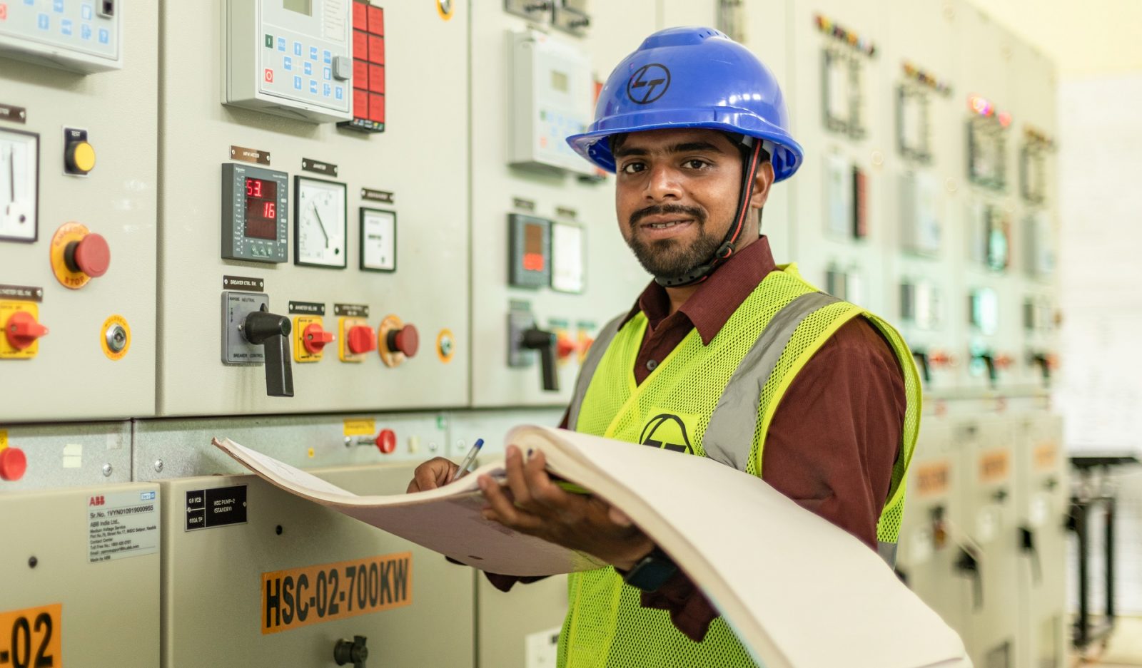 A man wearing a blue hard hat working at a machine while holding a large log book