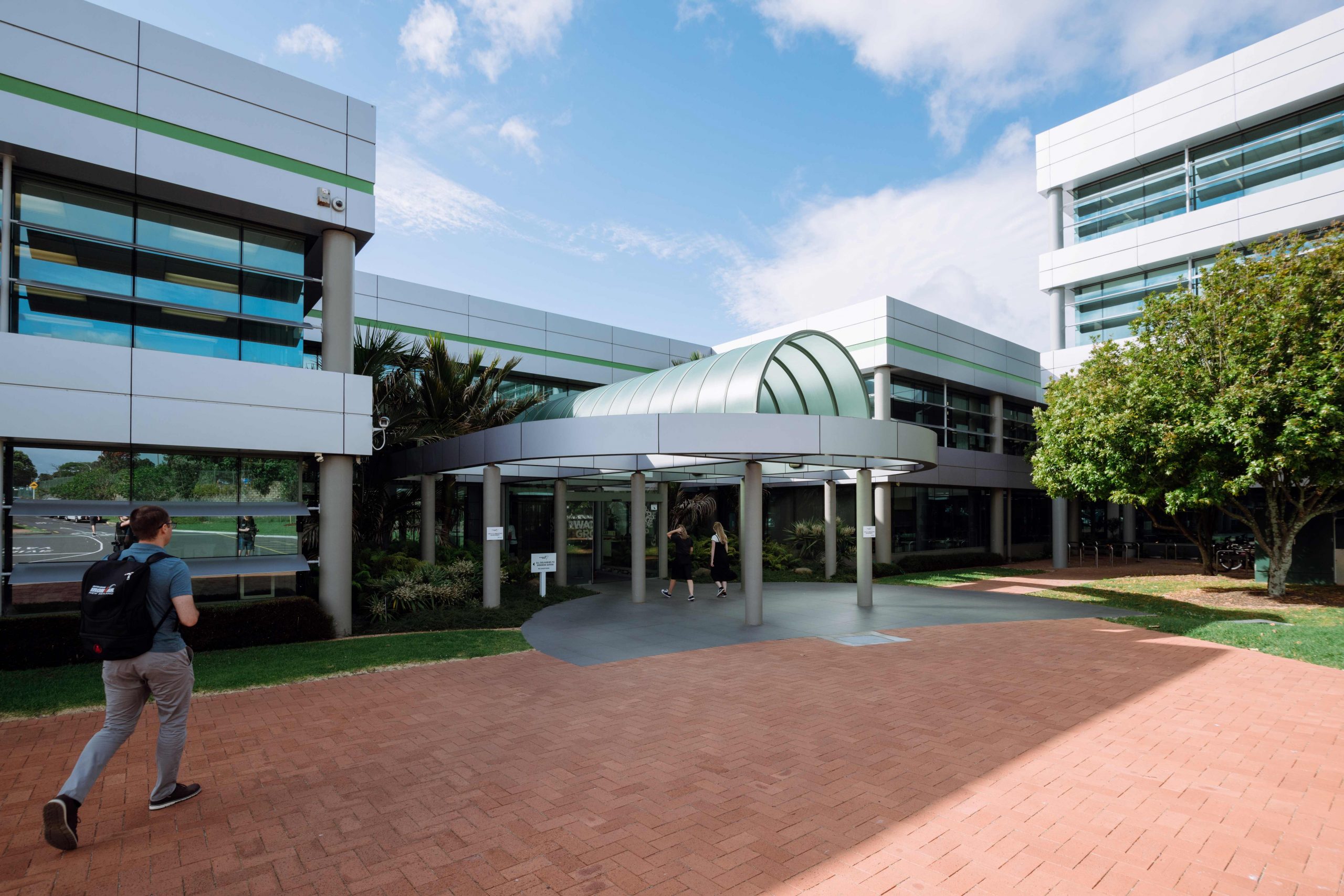 Exterior view of a modern office building with large glass windows and a central covered entrance. Two people walk towards the entrance, while another person is walking away, carrying a backpack. The building is surrounded by greenery and features a clear sky in the background.