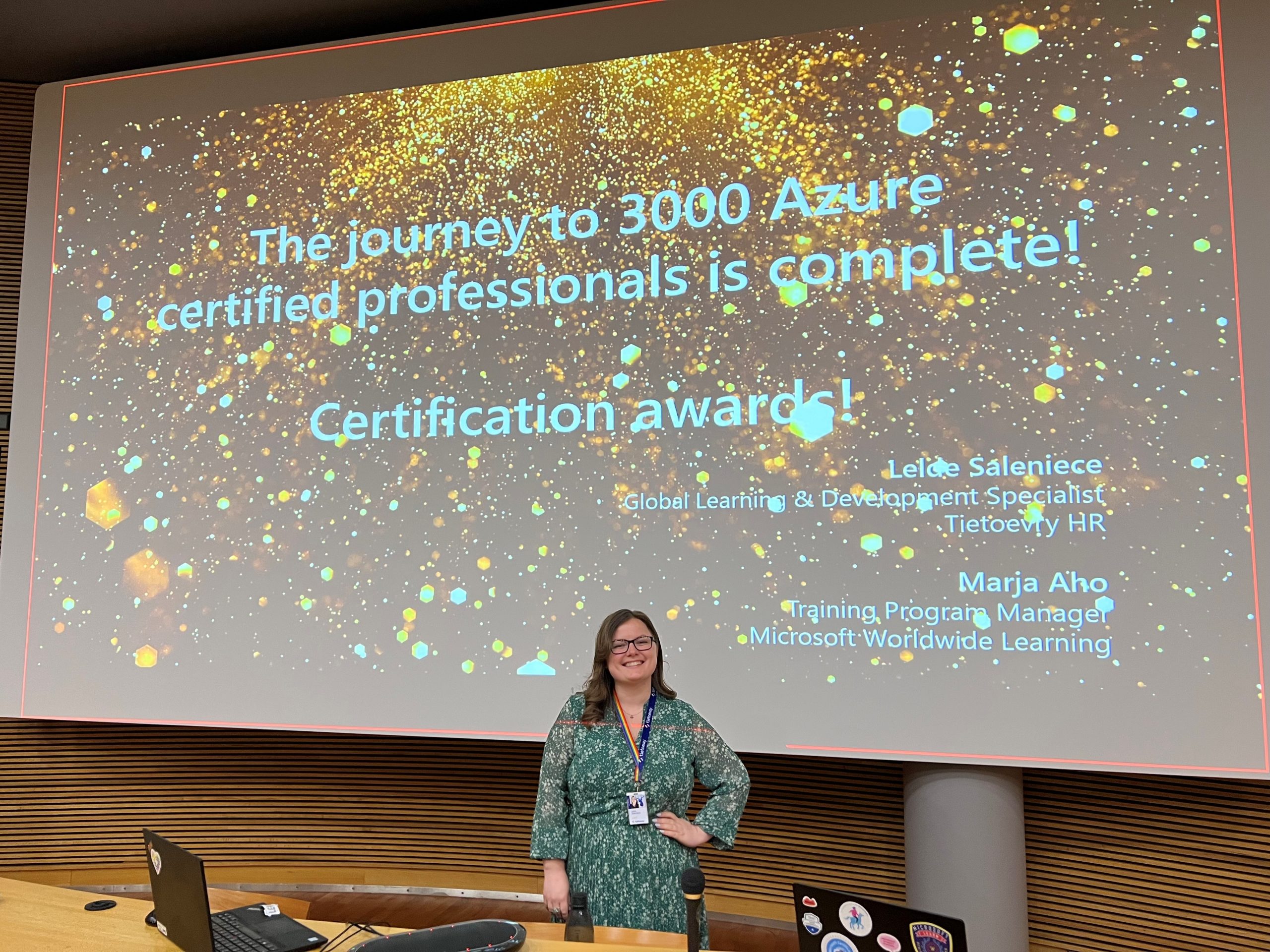 Nainen seisoo etualalla ja hymyilee. Taustalla teksti: "The journey to 3000 Azure certified professionals is complete! Certification awards".