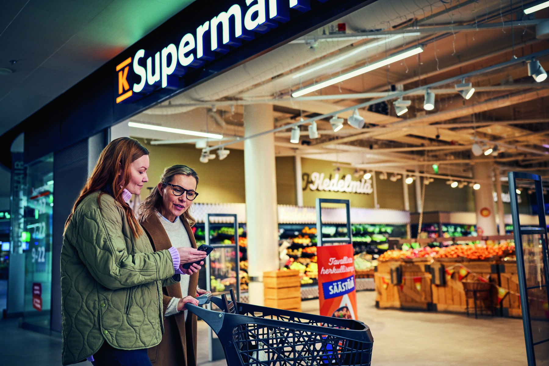 Kaksi naista tutkii puhelinta K-supermarketin edessä.