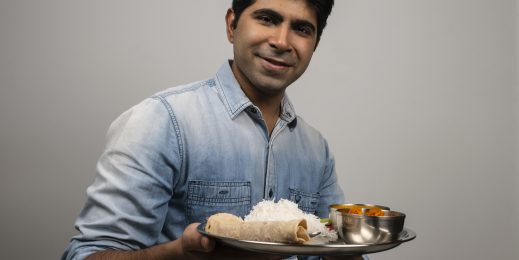 a man wearing a light blue denim shirt holding a plate of food in his hands and looking at the camera