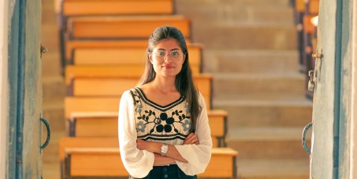 a woman wearing glasses and standing with her hands crossed in front of an empty classroom