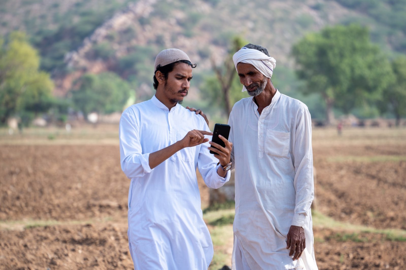 Two farmers standing in an arid village field looking at a mobile phone.