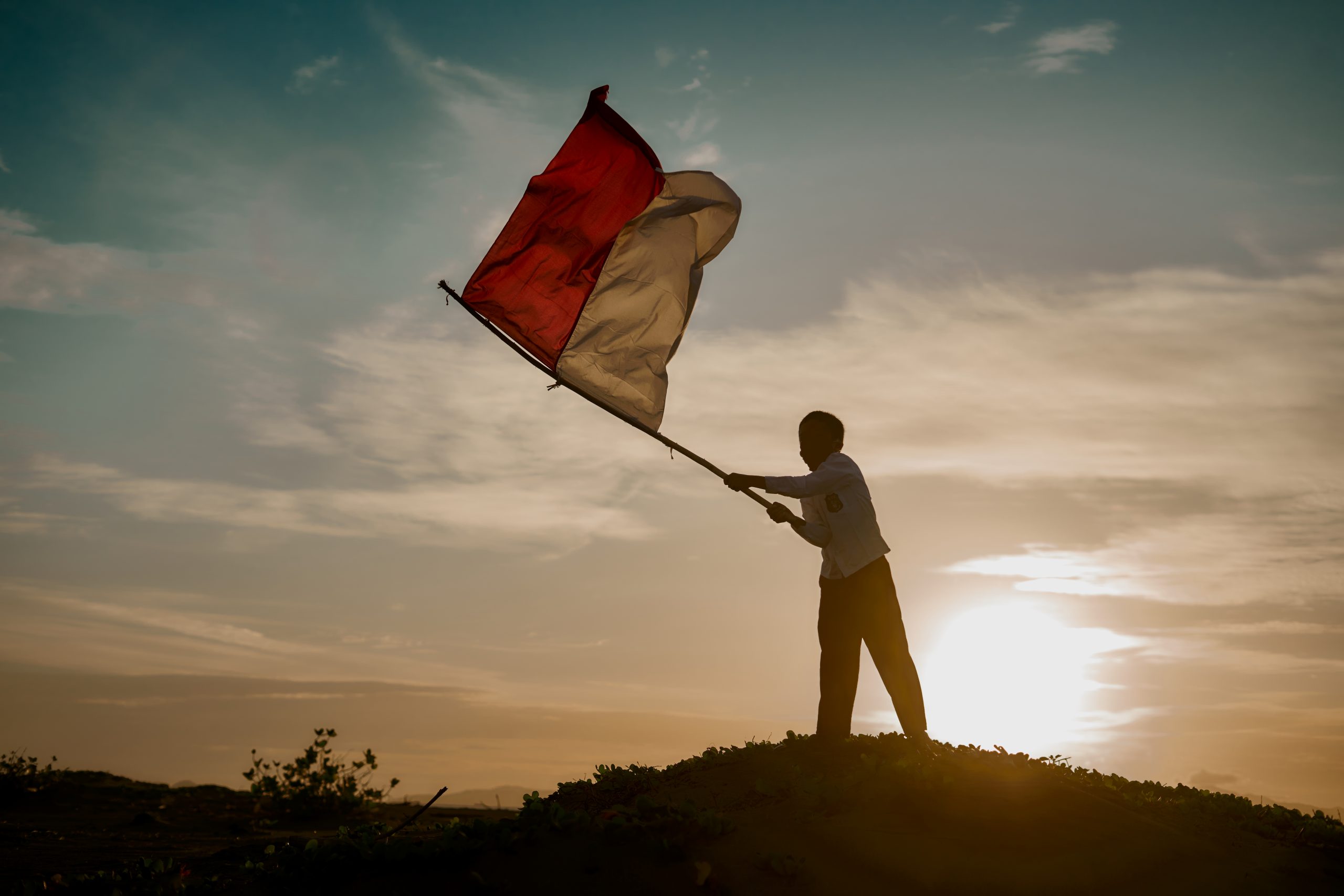 A photo of a student waving the Indonesian flag on a hill at sunset