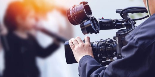 Руки оператора держат телекамеру, на заднем плане ведущая