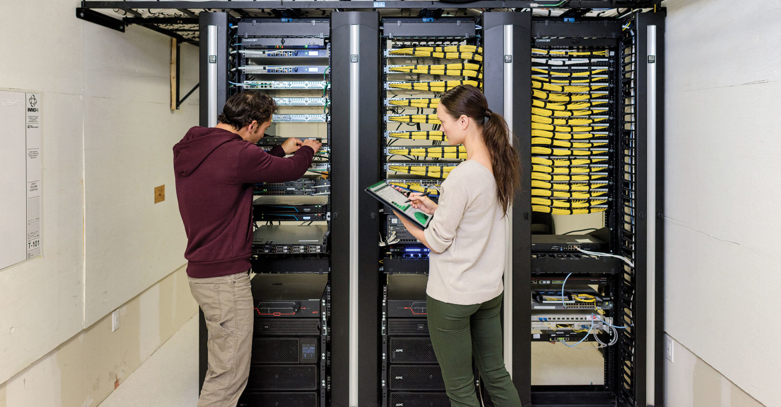 Женщина и мужчина стоят в серверной. Он что-то делает в стойке, она держит в руках планшет, на экране которого отображаются графики