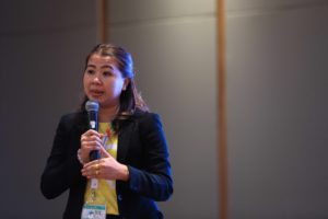 Woman in suit jacket and yellow shirt speaks on stage with microphone