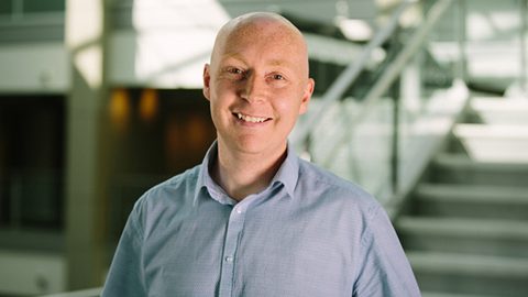Ryen White smiles into the camera in an office lobby, blurred staircase in the background