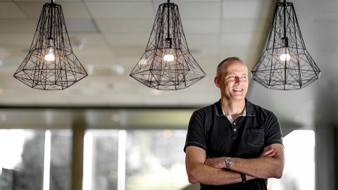 Todd Holmdahl stands with arm crossed, smiling off in the distance, with metal chandeliers in the background