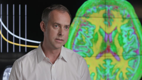 Mark Griswold talks to an interviewer in front of a screen showing MRI scans of a human brain