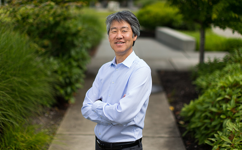 Peter Lee stands with arms crossed on a sidewalk surrounded by greenery