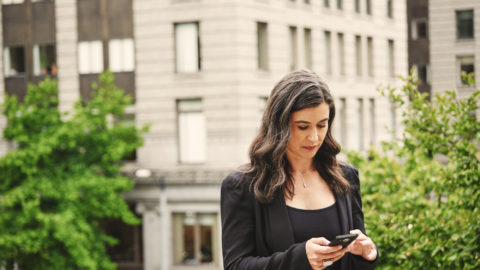 Businesswoman in blazer holds phone and types into it. Old city building makes up the entire background behind her.