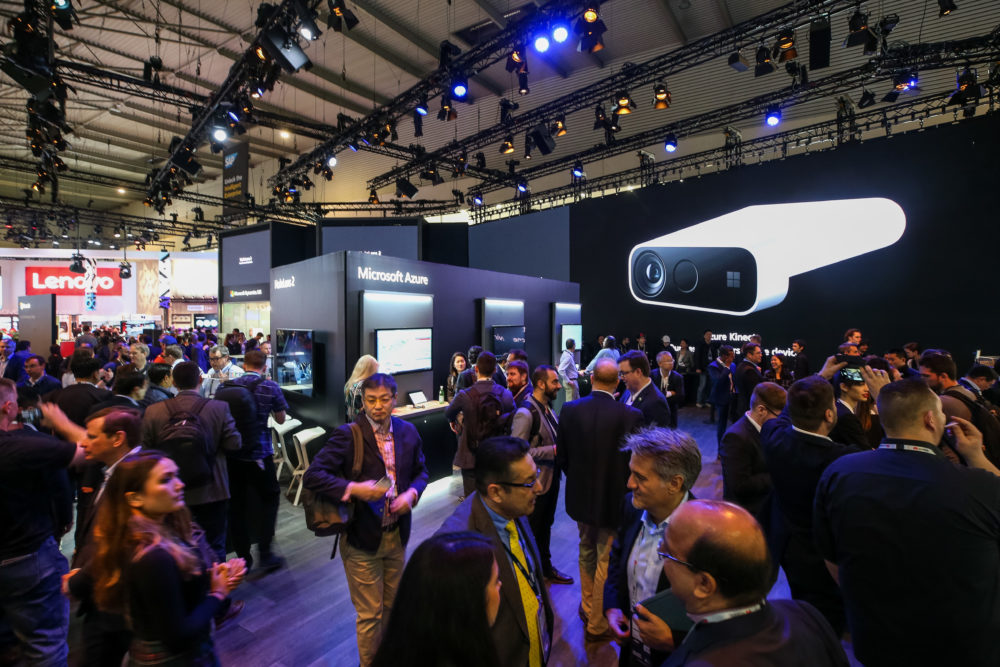 A large crowd of people mills around the Microsoft booth, where a large image of an Azure Kinect DK is displayed on one wall