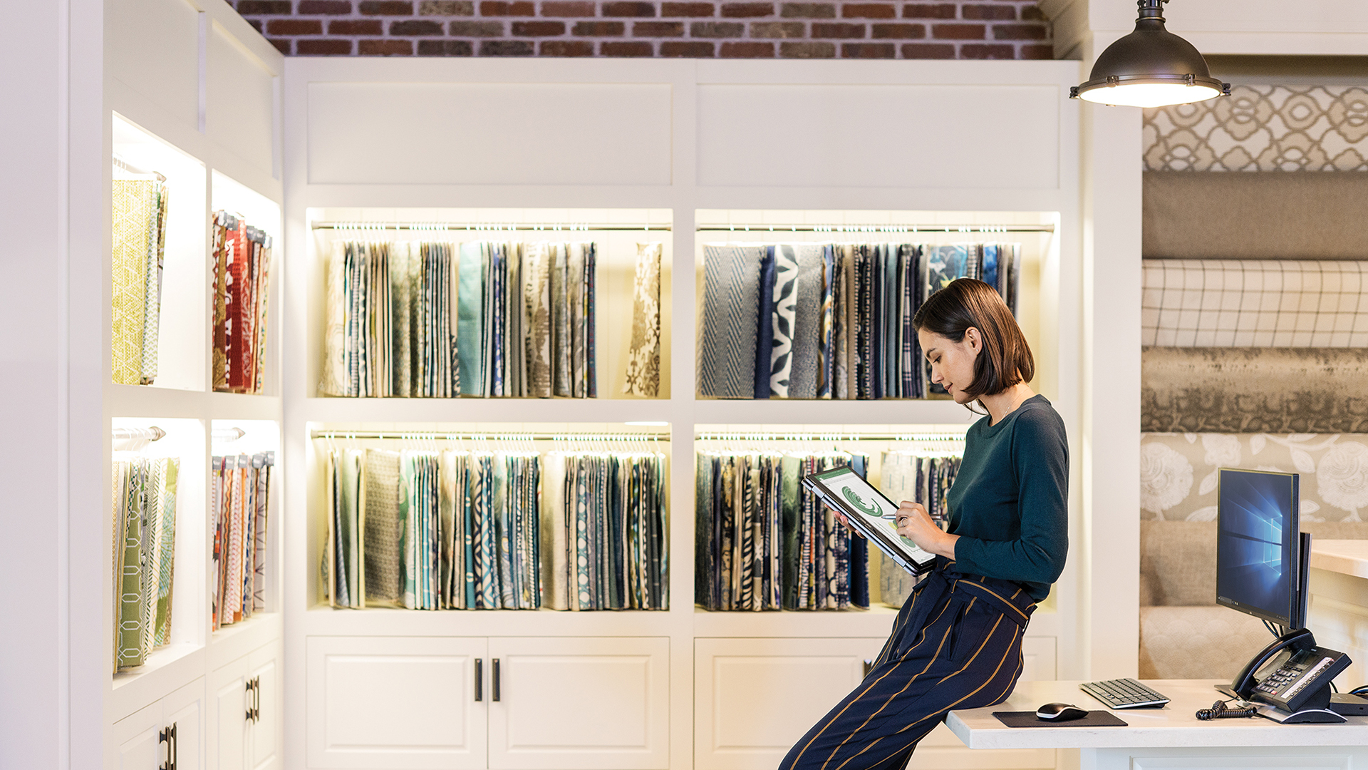A woman leans against a table holding a tablet, with a wall of tapestries beside her