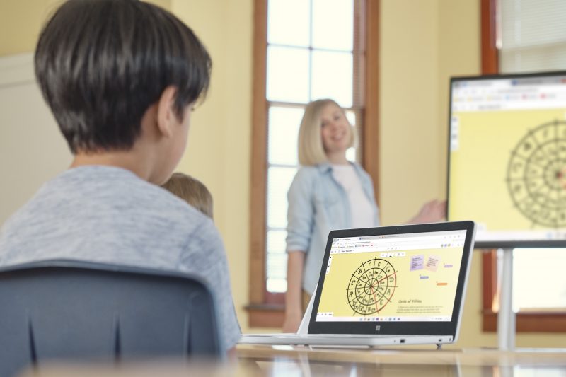 A boy sits in a classroom viewing his laptop as a teacher smiles